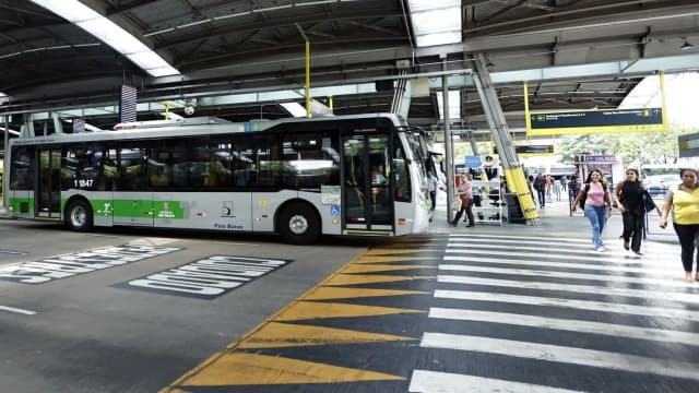 Ônibus do transporte público de São Paulo em terminal com passageiros passando pela faixa de pedestres