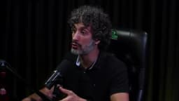 Carlos Eduardo Grimaldi gesticula com as mãos enquanto fala de frente ao microfone, em estúdio de podcast com cortina escura ao fundo