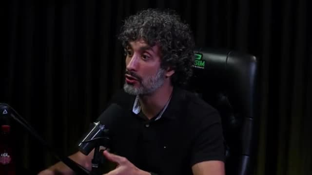 Carlos Eduardo Grimaldi gesticula com as mãos enquanto fala de frente ao microfone, em estúdio de podcast com cortina escura ao fundo