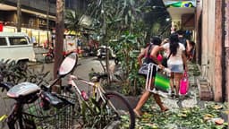 Pedestres caminham por calçada carioca coberta de folhas e galhos arrancados pela ventania, à noite, com bicicleta e moto encostadas e comércio ao fundo