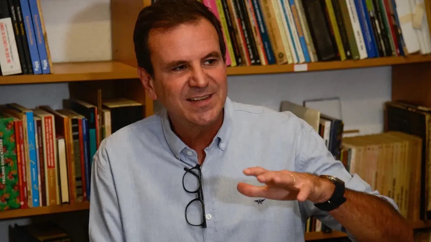 Eduardo Paes gesticula durante entrevista informal, em frente a uma estante de livros, vestindo camisa social azul clara
