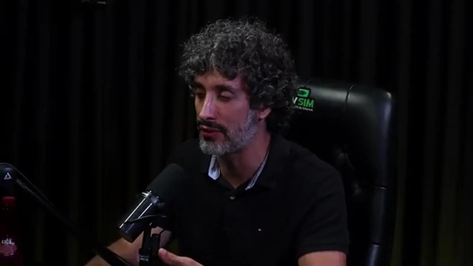 Carlos Eduardo Grimaldi fala olhando de lado ao microfone, com expressão séria, em estúdio de podcast com cortina escura ao fundo