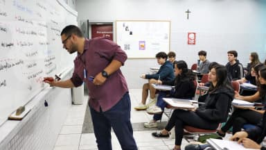 Professor escreve no quadro branco diante de uma turma sentada em carteiras