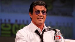 Sylvester Stallone usa óculos escuros durante evento público, sorrindo diante de microfone