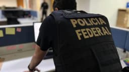 Agente da Polícia Federal com colete escrito Polícia Federal, visto de costas em escritório