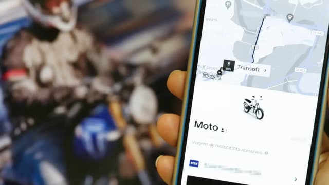 Mão segura celular com o aplicativo da Uber aberto na opção moto, com motociclista ao fundo