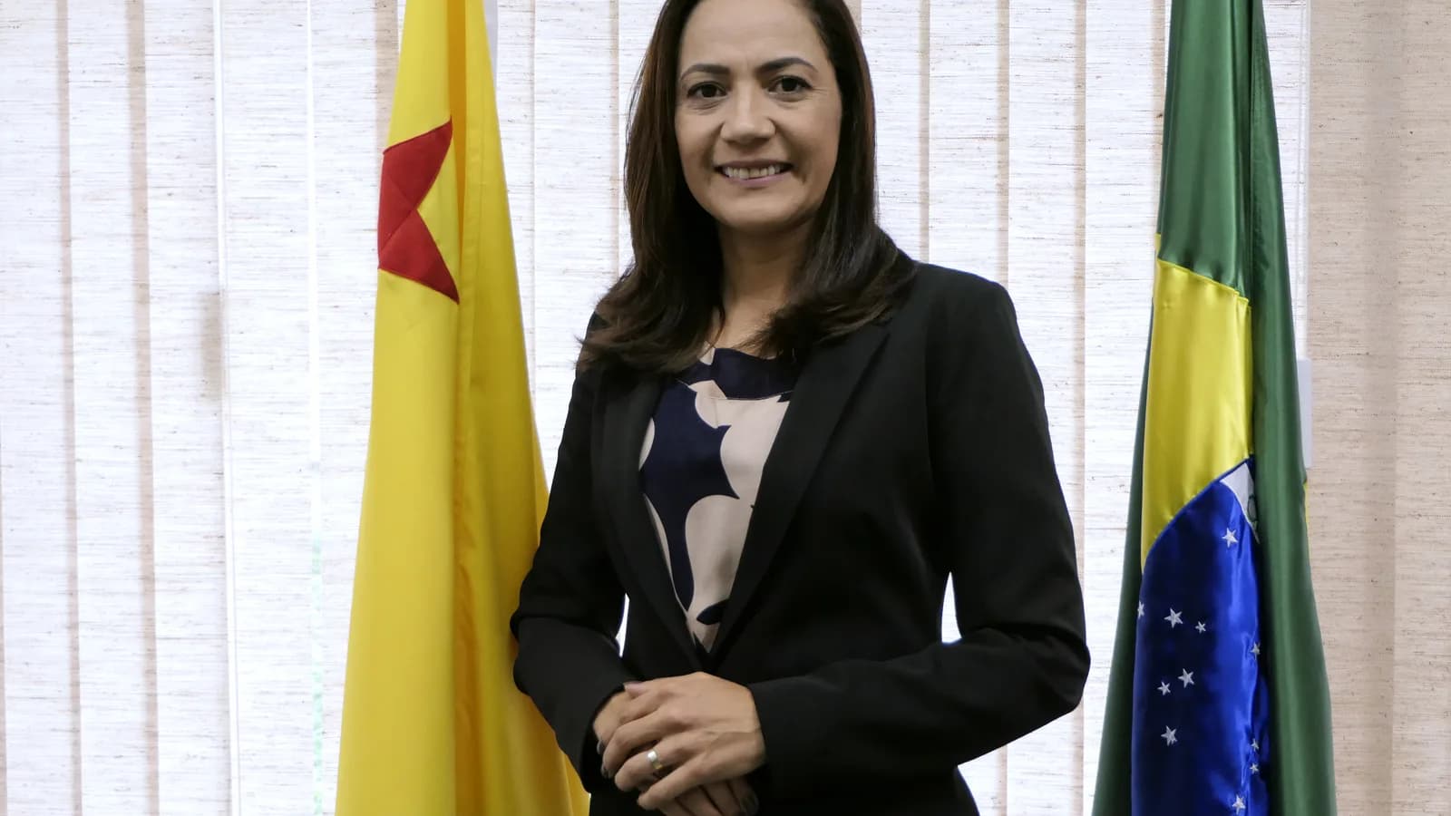 Mailza Assis posa em ambiente institucional, entre as bandeiras do Acre e do Brasil