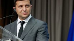 Volodymyr Zelensky fala ao microfone em pronunciamento, com bandeira da Ucrânia ao fundo