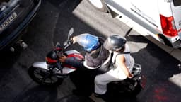 Vista de cima de uma motocicleta parada no trânsito entre carros, com piloto e passageiro usando capacete aberto; imagem de arquivo