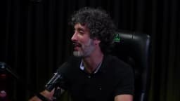 Carlos Eduardo Grimaldi sorri enquanto fala ao microfone, olhar erguido, em estúdio de podcast com cortina escura ao fundo