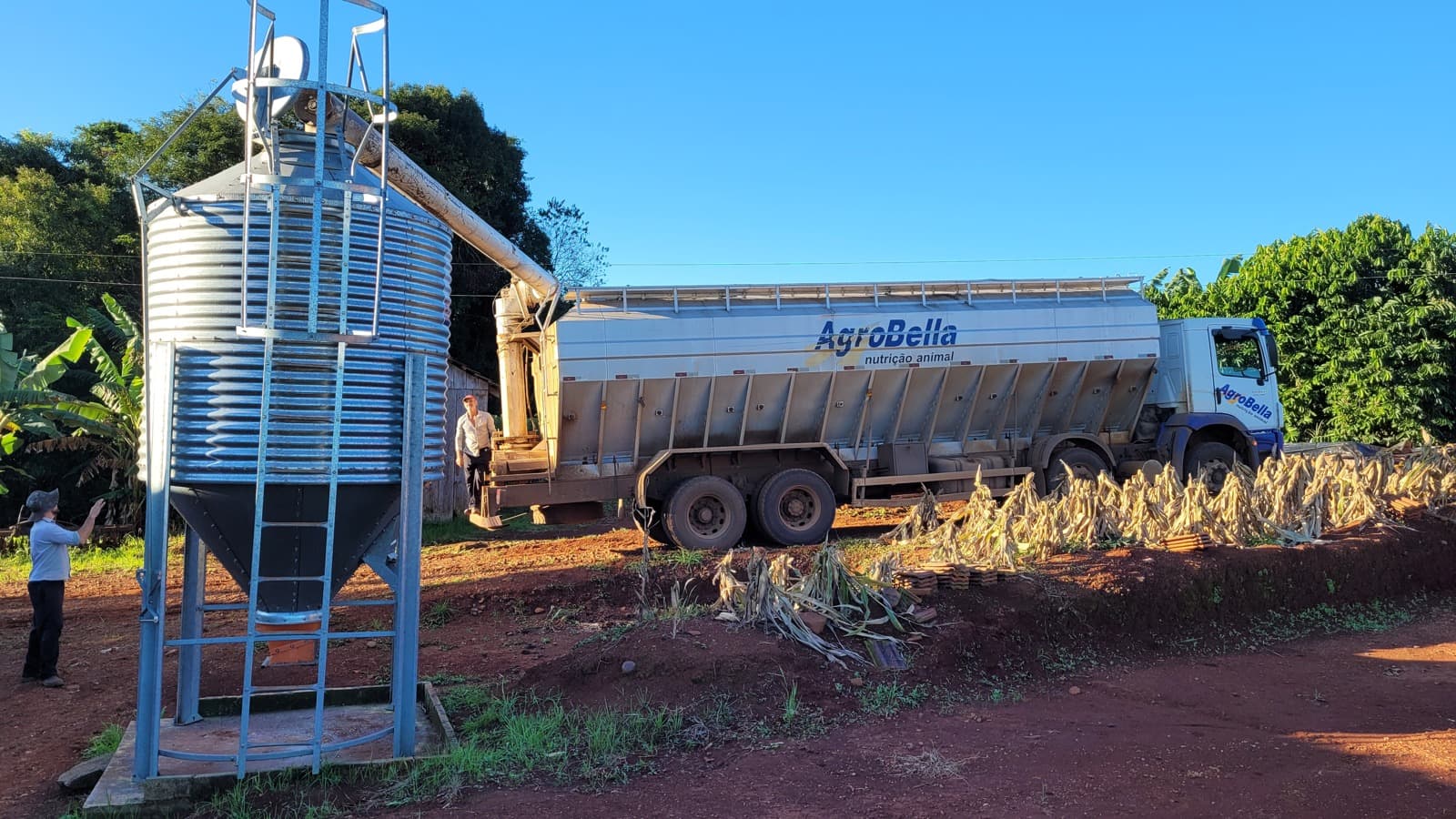 Caminhão de nutrição animal descarrega ração em silo vertical numa propriedade rural, com dois trabalhadores no local