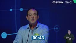 Flávio Roscoe, candidato ao governo de Minas Gerais, fala ao microfone no púlpito do debate, com telão azul ao fundo