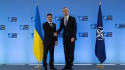 Volodymyr Zelensky aperta a mão de autoridade da Otan diante de bandeiras da Ucrânia e da aliança