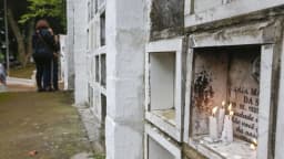 Pessoas em frente a gavetas de um cemitério em São Paulo, com velas acesas em memória de um ente querido