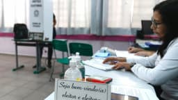 Mesária trabalha em mesa de votação com urna eletrônica ao fundo em seção eleitoral