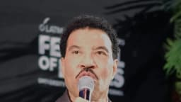O cantor Lionel Richie segura um microfone e fala durante evento, vestindo jaqueta de couro