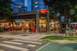 Fachada de esquina do Mercado da Boca ao anoitecer, com o logotipo de boca vermelha no alto do prédio, as operações Trip's Burger e Esquina iluminadas e clientes sentados nas mesas da calçada.