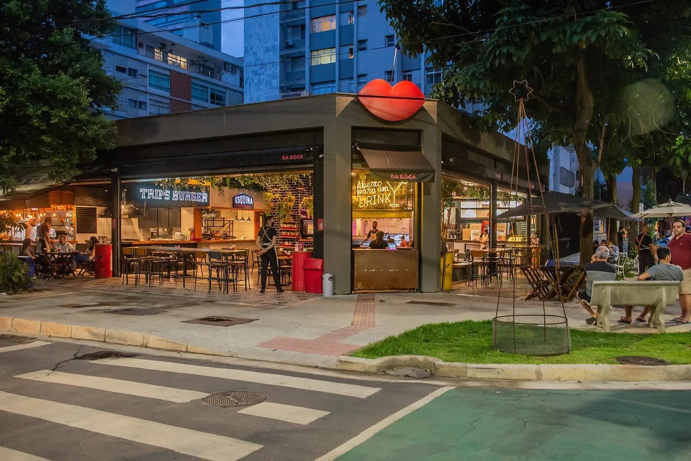 Fachada de esquina do Mercado da Boca ao anoitecer, com o logotipo de boca vermelha no alto do prédio, as operações Trip's Burger e Esquina iluminadas e clientes sentados nas mesas da calçada.
