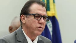 O ministro do Trabalho e Emprego, Luiz Marinho, falando ao microfone diante da bandeira do Brasil