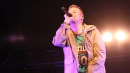 Macklemore canta ao microfone durante show, com luzes de palco ao fundo