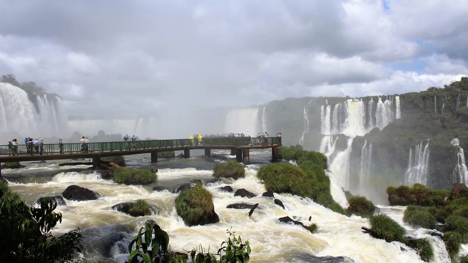 Visitantes caminham pela passarela das Cataratas do Iguaçu em meio a água turva e volume elevado do rio