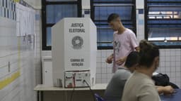 Eleitor se aproxima de uma cabina de votação da Justiça Eleitoral em uma seção eleitoral, com mesários à mesa; imagem de arquivo de pleito anterior