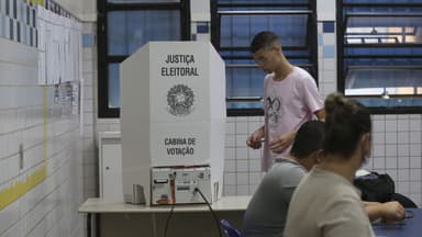 Eleitor se aproxima de uma cabina de votação da Justiça Eleitoral em uma seção eleitoral, com mesários à mesa; imagem de arquivo de pleito anterior