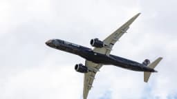Avião Embraer E195-E2 em voo, visto de baixo contra o céu com nuvens