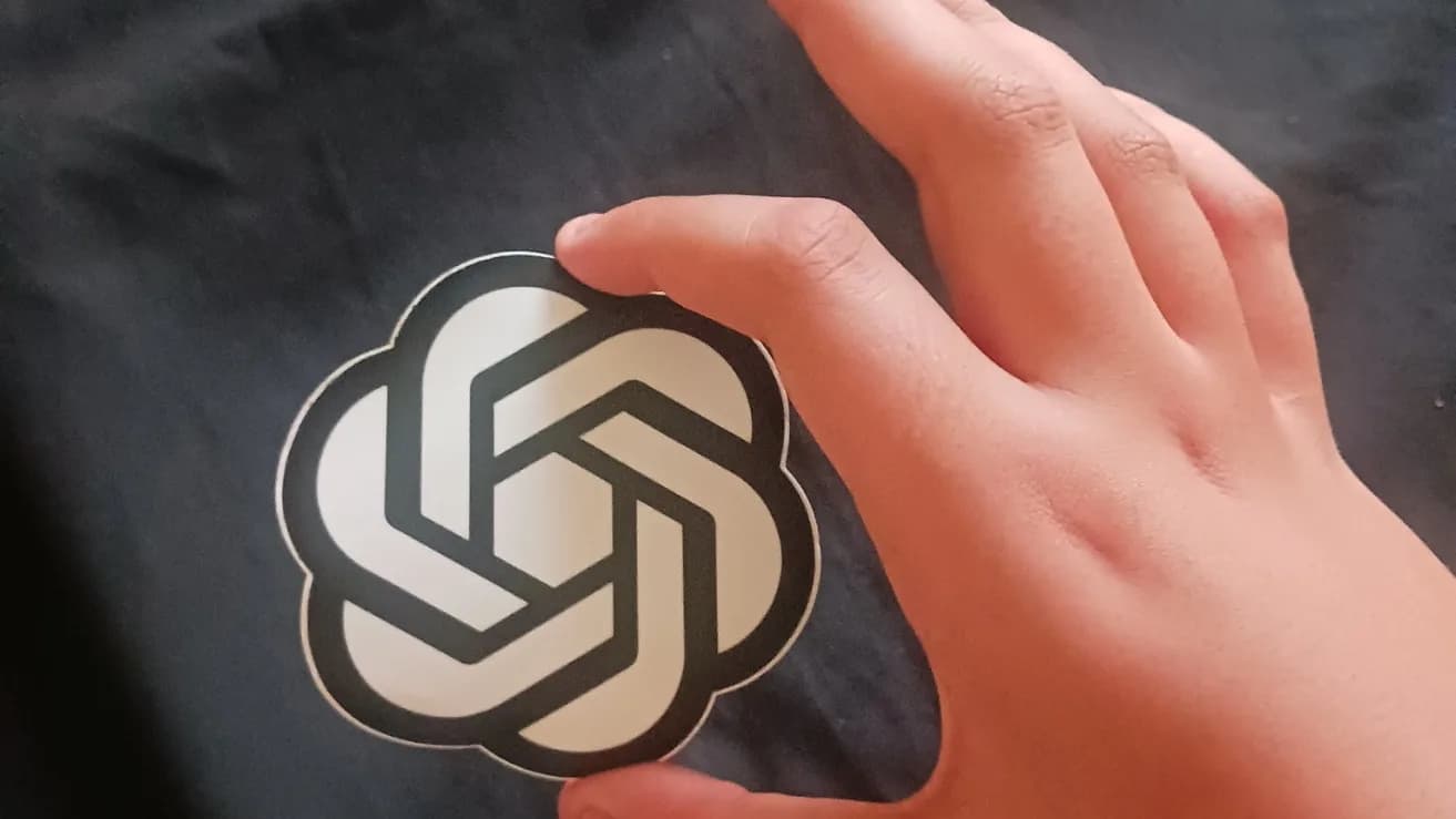 Mão segurando um adesivo circular com o logo da OpenAI