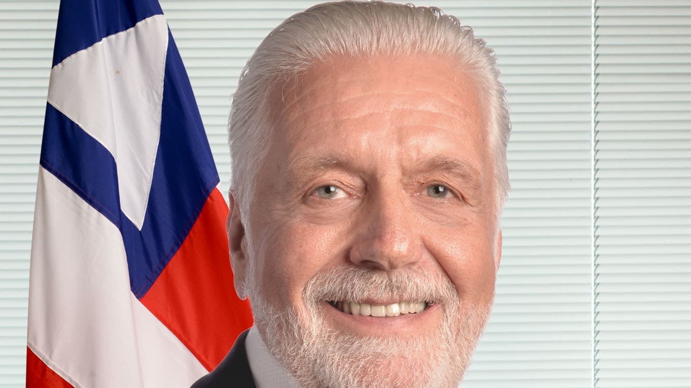 Retrato oficial do senador Jaques Wagner, de terno escuro e gravata vermelha, com a bandeira da Bahia ao fundo