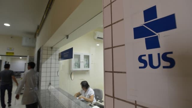Logo do SUS na parede de corredor de unidade de saúde, com profissional de jaleco e atendente ao fundo (foto de arquivo)
