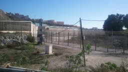 Cerca dupla de arame com torre de vigilância na fronteira entre Ceuta e o Marrocos; ao fundo, prédios da cidade em uma encosta. Imagem de arquivo.