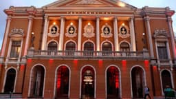 Fachada iluminada do Theatro da Paz, em Belém, com colunas, arcos e o nome do teatro no alto do frontão ao entardecer