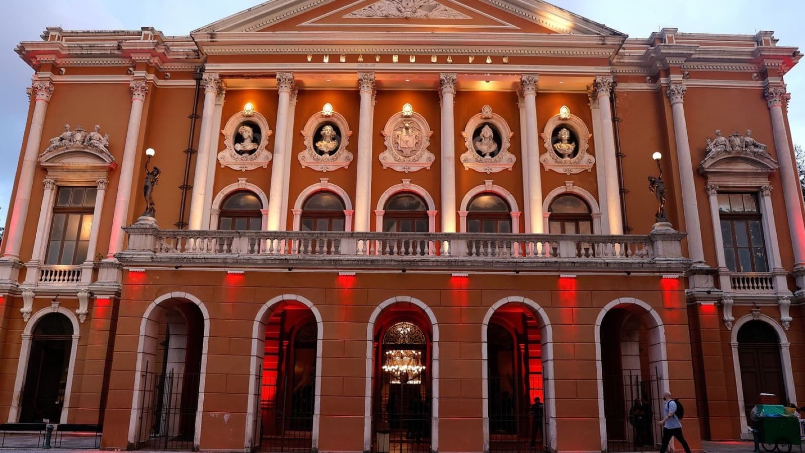 Fachada iluminada do Theatro da Paz, em Belém, com colunas, arcos e o nome do teatro no alto do frontão ao entardecer