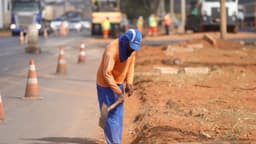 Trabalhador de uniforme laranja e azul e boné cava com uma enxada na beira de uma obra de recuperação de rodovia no Distrito Federal, com cones e caminhões ao fundo; foto de arquivo, ilustrativa