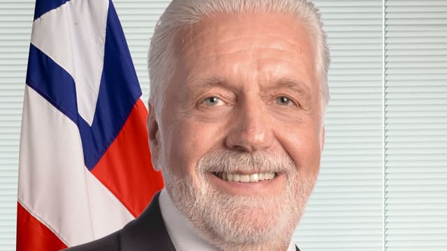Retrato oficial do senador Jaques Wagner, de cabelos e barba brancos e terno escuro, sorrindo diante da bandeira da Bahia