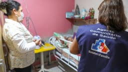 Agente comunitária de saúde do SUS em visita domiciliar, com colete identificado 'Agente Comunitário de Saúde - Saúde da Família'