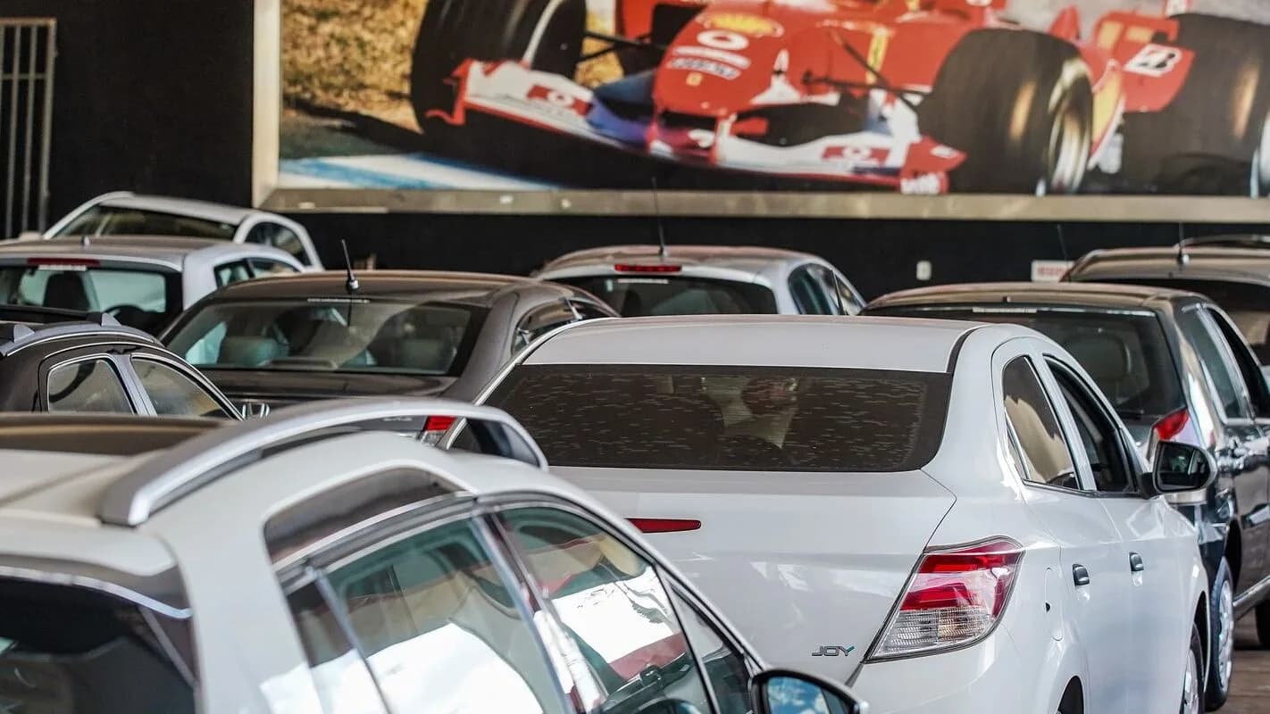 Pátio com veículos novos estacionados para exportação, com pintura de carro de corrida ao fundo