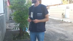 Agente do IBGE com colete oficial e crachá caminha por rua residencial em trabalho de campo