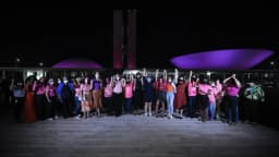 Grupo de pessoas reunido em evento de conscientização do Outubro Rosa, com as cúpulas do Congresso Nacional iluminadas de rosa ao fundo