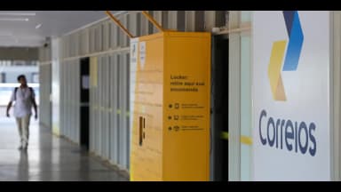 Locker dos Correios para retirada de encomendas em corredor de prédio público, com pessoa caminhando ao fundo