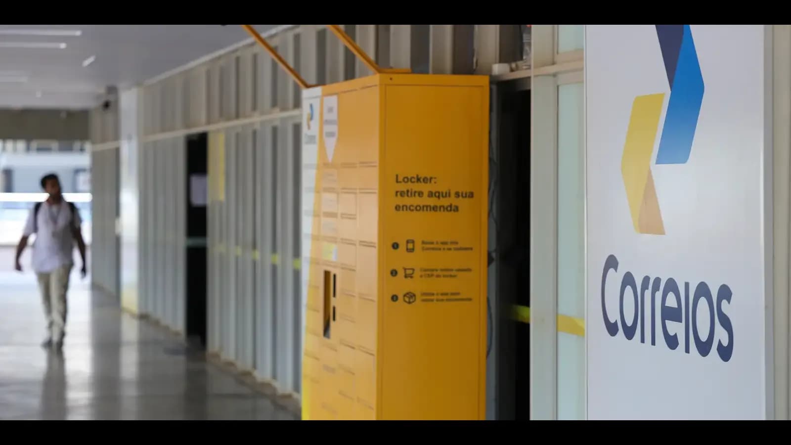 Locker dos Correios para retirada de encomendas em corredor de prédio público, com pessoa caminhando ao fundo