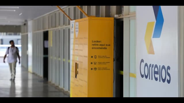 Locker dos Correios para retirada de encomendas em corredor de prédio público, com pessoa caminhando ao fundo
