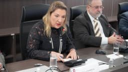 Daniella Marques, de blusa estampada e colar de pérolas, fala ao microfone em mesa de reunião