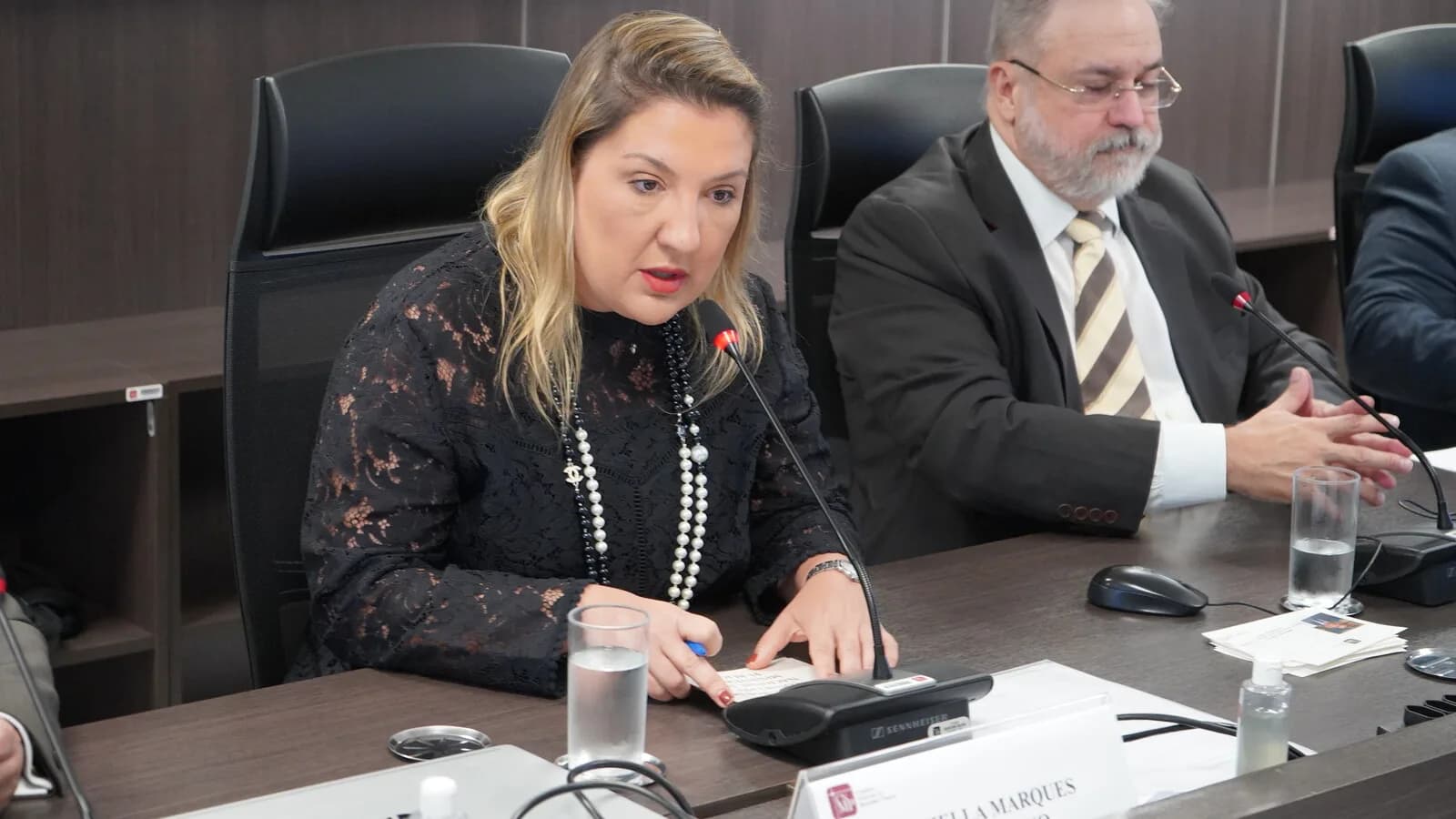 Daniella Marques, de blusa estampada e colar de pérolas, fala ao microfone em mesa de reunião
