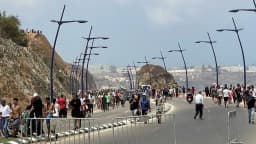 Multidão caminha por avenida à beira-mar em Ceuta, com postes de iluminação ao longo da via