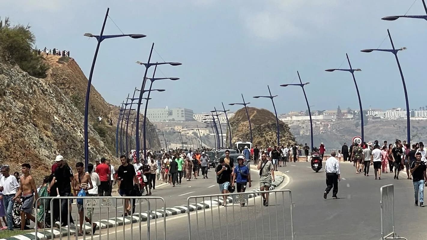 Multidão caminha por avenida à beira-mar em Ceuta, com postes de iluminação ao longo da via