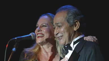 Gracindo Jr. sorri ao lado da atriz Lucinha Lins, em cena de espetáculo musical, os dois vestidos a rigor diante de um microfone