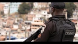 Policial militar de costas, com colete tático escrito Polícia, observa comunidade em morro no Rio de Janeiro