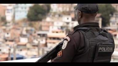 Policial militar de costas, com colete tático escrito Polícia, observa comunidade em morro no Rio de Janeiro